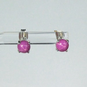 May include: A pair of stud earrings with round, magenta gemstones displaying a star effect. The earrings have a silver metal setting with four prongs securing each stone. The background is a light, neutral colour.