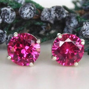 May include: A pair of round-cut, vibrant fuchsia-pink stud earrings. Each gemstone is secured in a four-prong silver-toned metal setting, showcasing a faceted surface that reflects light with a brilliant, deep pink color and clear clarity.