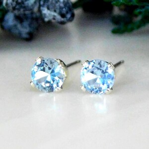 May include: A pair of round, light blue stud earrings featuring faceted stones set in four-prong metal settings. The earrings have a brilliant cut, reflecting light with a clear, icy blue hue, and include thin metal posts extending from the back.