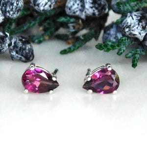 May include: A pair of stud earrings featuring pear-shaped, faceted magenta gemstones set in silver-toned metal prongs. The earrings are positioned side-by-side, showcasing the deep pink color and the reflective, multi-faceted surface of the stones.