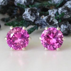 May include: A pair of stud earrings featuring large, round, faceted pink gemstones set in silver-colored metal. The earrings are displayed on a white surface with a blurred background of greenery and dark berries.