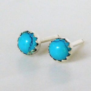 May include: A pair of stud earrings featuring round, bright turquoise stones with dark matrix veining. Each stone is set in a scalloped silver-toned metal bezel with a straight post extending from the back, designed for pierced ears.