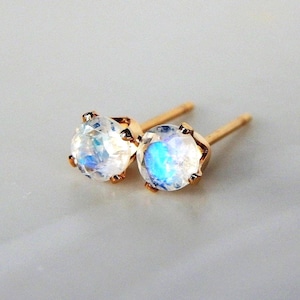 May include: A pair of stud earrings featuring round, faceted moonstones set in gold-toned metal prongs. Each earring has a straight post back. The moonstones exhibit a translucent appearance with a distinct blue iridescent glow visible.