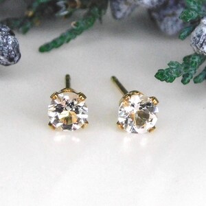May include: A pair of stud earrings featuring round, clear, faceted stones set in gold-toned metal prongs. Each earring has a straight post extending from the back, designed for pierced ears, showcasing a classic, reflective, and transparent appearance.