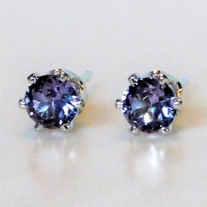 May include: A pair of stud earrings featuring round, faceted purple gemstones set in silver-toned metal prongs. Each earring has a circular cut stone that displays deep violet and blue hues, secured by a six-prong setting on a post back.