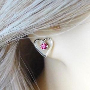 May include: A silver-toned heart-shaped stud earring featuring a small, round, vibrant pink gemstone set in the center. The metal frame forms a clean, open heart outline that surrounds the central stone, creating a simple and elegant design.