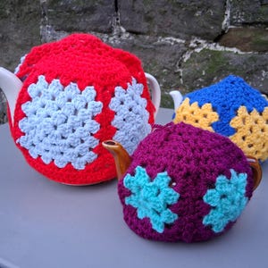 Può includere: Tre teiere con colorate coperture per tè all'uncinetto. Le coperture sono rosse con disegni floreali azzurri, blu con fiori gialli e viola con fiori turchesi. Le teiere sono bianche con beccucci e manici marroni.