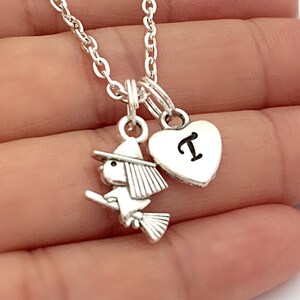 Puede incluir: Un collar plateado con dos colgantes: una bruja en una escoba y un corazón con la letra "T". El dije de la bruja tiene un sombrero y una escoba. El dije de corazón es liso y redondeado.