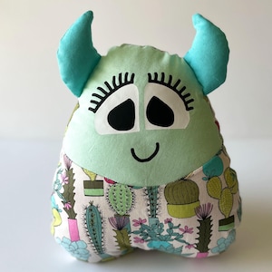 May include: A plush monster doll with a light green face, blue horns, and a patterned body featuring cacti. The doll has large black eyes, long eyelashes, and a smiling mouth. The soft toy is designed to be a decorative item.