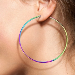 May include: A close-up of a person's ear wearing a large, iridescent hoop earring. The earring transitions through a spectrum of colors, including green, blue, and purple. The earring is a simple, circular design, and the focus is on the vibrant color.