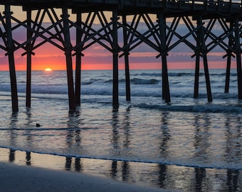 May include: A wooden pier extends over a calm ocean at sunrise. The sun is rising over the horizon, casting a warm glow on the water. The pier is made of dark wood and has many supports. The water is a deep blue color and is reflecting the light of the sun.