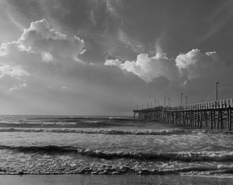 May include: A black and white photograph of a wooden pier extending out over a choppy ocean. The pier has several lights and is empty. The sky is cloudy and overcast.