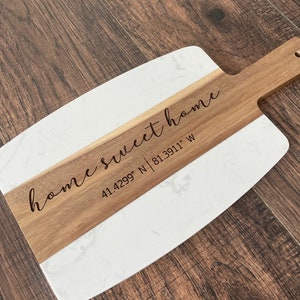 May include: Wooden serving board with a white marble-look section and a handle. The words "home sweet home" and coordinates 41.4299° N 81.3911° W are engraved on the wood. The board is on a dark wood surface.