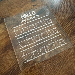 May include: A clear plastic learning board with the text "HELLO my name is" and the name "Charlie" written multiple times in dotted lines for tracing.
