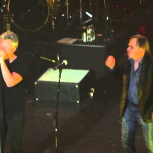May include: Two men on stage performing in front of a dark background. The man on the left is holding a microphone and singing. The man on the right is wearing a dark jacket and is gesturing with his right hand.