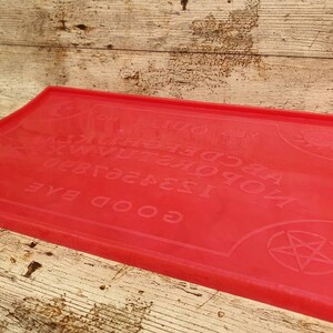 Puede incluir: Tablero de Ouija de silicona rojo con las letras A-Z, los n&uacute;meros 1-9 y las palabras "GOOD BYE", "YES", "NO" y "GOODBYE". El tablero tambi&eacute;n presenta un sol y un pentagrama.