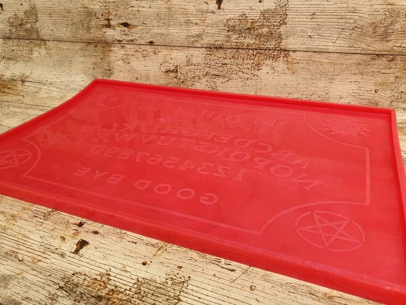 Puede incluir: Tablero de Ouija de silicona rojo con las letras A-Z, los n&uacute;meros 1-9 y las palabras "GOOD BYE", "YES", "NO" y "GOODBYE". El tablero tambi&eacute;n presenta un sol y un pentagrama.