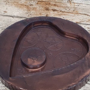 Puede incluir: Una bandeja de madera de color marr&oacute;n oscuro en forma de coraz&oacute;n con un dise&ntilde;o en relieve de un pentagrama, una luna creciente, el sol y otros s&iacute;mbolos. La bandeja tiene una hendidura circular en el centro.