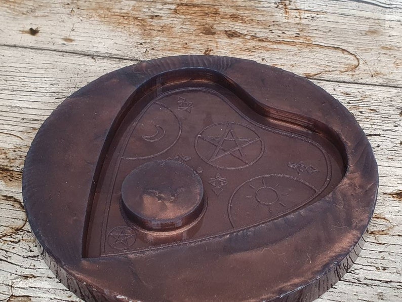 Puede incluir: Una bandeja de madera de color marr&oacute;n oscuro en forma de coraz&oacute;n con un dise&ntilde;o en relieve de un pentagrama, una luna creciente, el sol y otros s&iacute;mbolos. La bandeja tiene una hendidura circular en el centro.