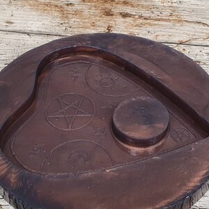 Puede incluir: Una tabla de Ouija de madera marr&oacute;n oscuro con un recorte en forma de coraz&oacute;n y una hendidura circular en el centro. La tabla est&aacute; decorada con s&iacute;mbolos que incluyen un pentagrama, una luna creciente y estrellas.