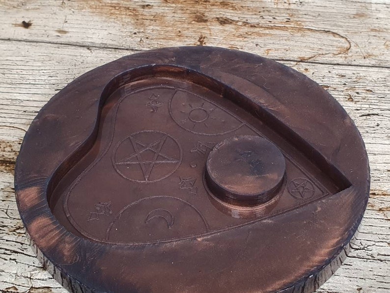 Puede incluir: Una tabla de Ouija de madera marr&oacute;n oscuro con un recorte en forma de coraz&oacute;n y una hendidura circular en el centro. La tabla est&aacute; decorada con s&iacute;mbolos que incluyen un pentagrama, una luna creciente y estrellas.