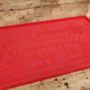 Puede incluir: Una tabla de Ouija roja con un pentagrama negro en cada esquina. La tabla tiene las letras del alfabeto, los n&uacute;meros del 0 al 9, y las palabras "s&iacute;", "no", "adi&oacute;s", y "buenas noches".