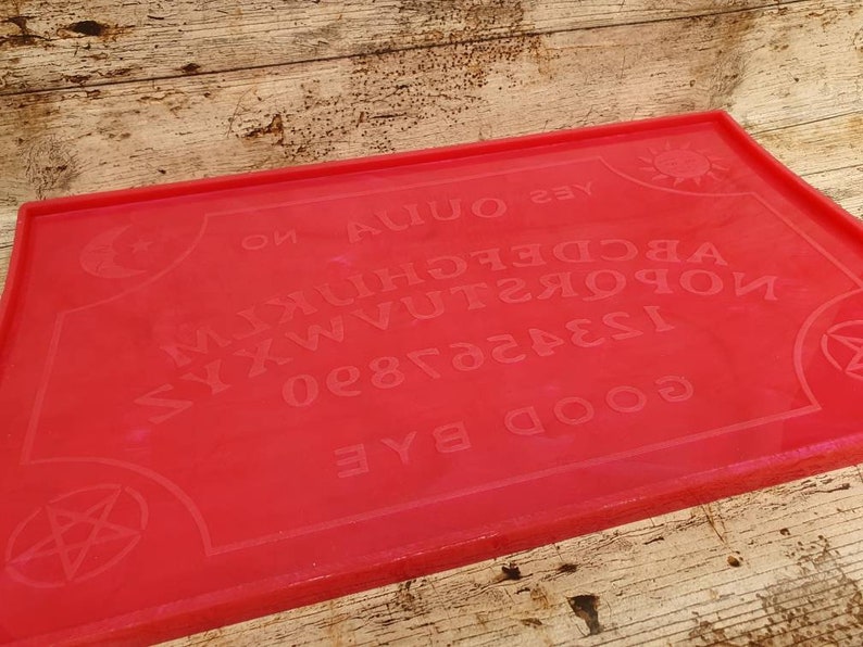 Puede incluir: Una tabla de Ouija roja con un pentagrama negro en cada esquina. La tabla tiene las letras del alfabeto, los n&uacute;meros del 0 al 9, y las palabras "s&iacute;", "no", "adi&oacute;s", y "buenas noches".