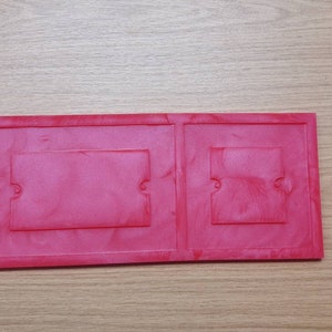 May include: A red plastic frame with two rectangular openings. The frame is made of a smooth, glossy plastic.