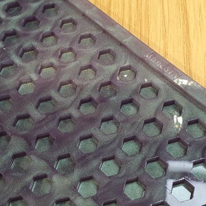 Puede incluir: Primer plano de una superficie texturizada de color morado oscuro y verde con un patrón de panal de abeja. La superficie tiene un ligero brillo y está cubierta por una fina capa de polvo. El texto "Made in China" es visible en la esquina superior derecha.
