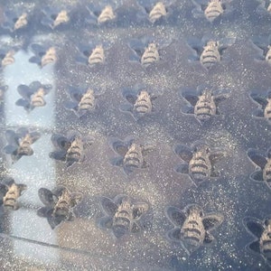 以下が含まれることがあります： 透明なガラスの表面をクローズアップした写真。ガラスに刻まれた蜂の形が繰り返しパターンを描いています。蜂の形は薄い茶色の物質で満たされています。表面は細かいほこりで覆われています。