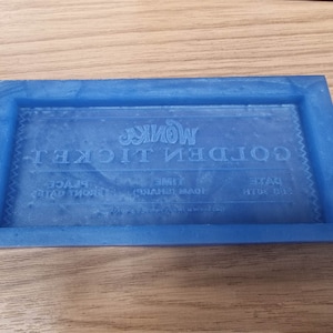 May include: A blue resin tray with a raised border, rectangular in shape. The tray features the words "GOLDEN TICKET" and "WONKA" in a stylized font, along with other text. It sits on a light brown wooden surface.