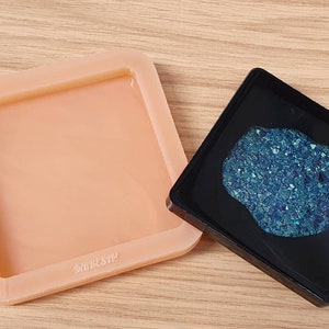 May include: A peach-colored square silicone mold with rounded corners, next to a black square tray containing a blue, crystalline substance. The mold has the text "AMRESYN" on the side. The items are on a light wood surface.