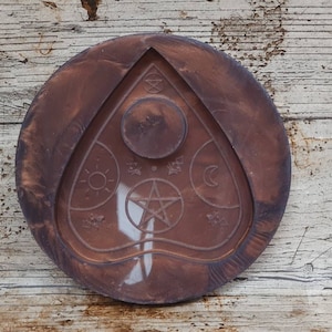 Puede incluir: Una bandeja redonda de madera marr&oacute;n con un dise&ntilde;o blanco en relieve de una planchette de tablero Ouija. La planchette presenta una estrella de cinco puntas, un sol, una luna y otros s&iacute;mbolos.