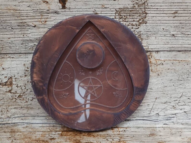 Puede incluir: Una bandeja redonda de madera marr&oacute;n con un dise&ntilde;o blanco en relieve de una planchette de tablero Ouija. La planchette presenta una estrella de cinco puntas, un sol, una luna y otros s&iacute;mbolos.