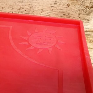 Puede incluir: Bandeja de silicona roja con un dise&ntilde;o de sol en relieve. El sol tiene una cara con un ce&ntilde;o fruncido.