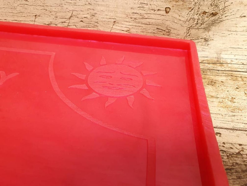 Puede incluir: Bandeja de silicona roja con un dise&ntilde;o de sol en relieve. El sol tiene una cara con un ce&ntilde;o fruncido.