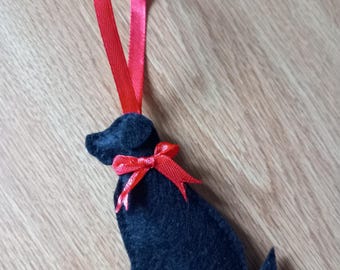 Può includere: Ornamento a forma di cane in feltro nero con fiocco di nastro rosso e anello rosso per appenderlo. Il cane è seduto. L'ornamento è su una superficie di legno.