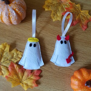 May include: Two felt ghost decorations, one with a halo and one with horns, hanging from white ribbon. The ghosts are white with black eyes and mouths. The halo is yellow and the horns are red. The decorations are surrounded by autumn leaves and pumpkins.
