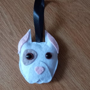 May include: A white felt dog ornament with pink ears and a brown nose. The dog has black button eyes and a black ribbon for hanging.