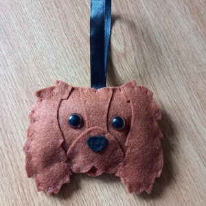 May include: A brown felt dog decoration with black button eyes and a black nose. The dog has floppy ears and a black satin ribbon for hanging. The ornament is on a wooden surface.