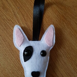 May include: A felt Bull Terrier dog head ornament. The dog is white with a black eye patch and nose, and pink ears. It has black button eyes and a black ribbon for hanging. The ornament is handmade.