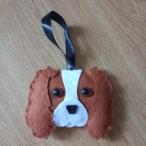 May include: A felt ornament shaped like a Cavalier King Charles Spaniel dog's head. The ornament is brown, white and black with black eyes and a black nose. It has a black ribbon for hanging.