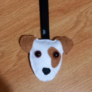 May include: A handmade dog-shaped keychain. The keychain features a white felt face with brown patches around the eyes and ears, and a black nose. It has two dark brown button eyes and a black strap.