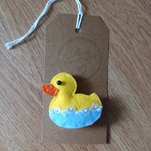 May include: A yellow felt rubber ducky with orange beak and black eyes, floating in blue felt water with white sequins for bubbles. The ducky is attached to a brown paper tag with a white string. The tag has a circle with the text "Made with love by" and "Jane Crafts" inside.