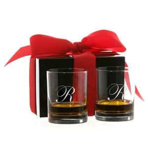 May include: Two clear whiskey glasses with the letter "R" etched on them, filled with amber liquid, sit in front of a black gift box tied with a red ribbon. The glasses are a classic rocks glass style.