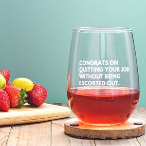 May include: A stemless wine glass filled with red liquid, resting on a wooden coaster. The glass has the text "CONGRATS ON QUITTING YOUR JOB WITHOUT BEING ESCORTED OUT." Strawberries and grapes sit on a wooden cutting board.