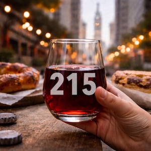 May include: A clear stemless wine glass filled with red wine, featuring the number "215" etched on the front. The glass is held by a hand, with a blurred background of a city scene and string lights. Bottle caps and food items are on the wooden table.