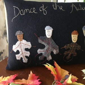 May include: A navy blue decorative pillow with the embroidered text "Dance of the Autumn". The pillow features appliqued leaf figures with smiling faces, connected by red thread, and a fall leaf garland.