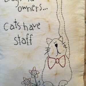 May include: Embroidered square cushion with the text "Dogs have owners... Cats have staff." A cartoon cat with a red bow tie is stitched on the cushion. Two embroidered flowers are near the cat.