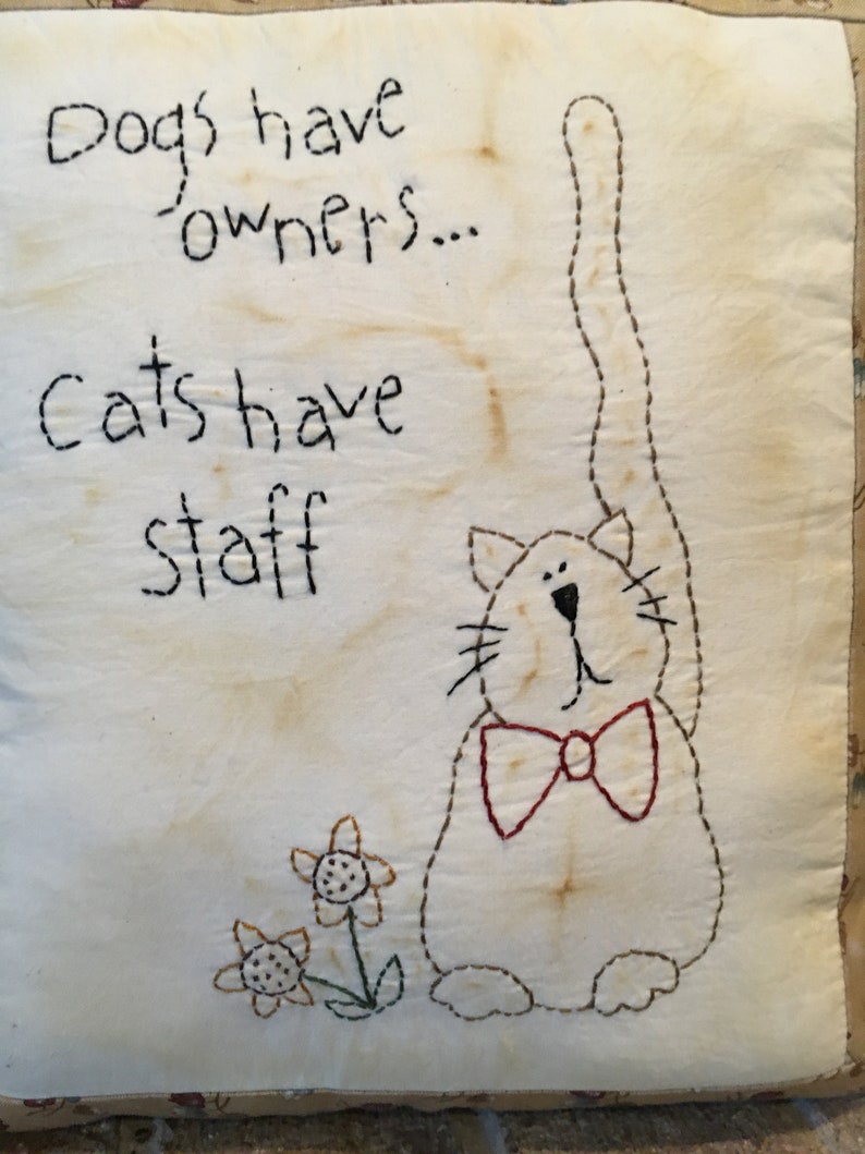 May include: Embroidered square cushion with the text "Dogs have owners... Cats have staff." A cartoon cat with a red bow tie is stitched on the cushion. Two embroidered flowers are near the cat.